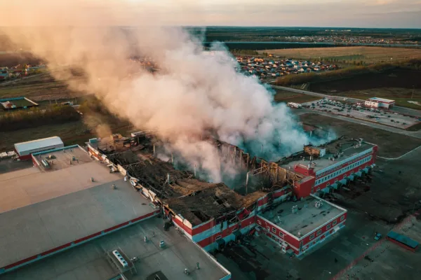 Aerial view of demolished burning industrial building by fire, huge smoke from broken roof, walls and piles of garbage